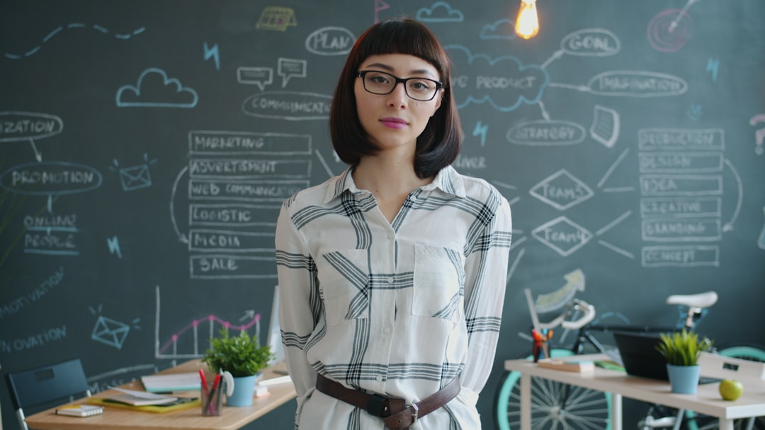 Portrait of attractive young lady standing in creative office with serious face and looking at camera. Modern youth, occupation and workspace concept.